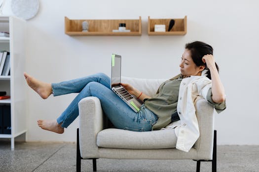 Asian woman lounging in an armchair, working remotely on a laptop at home.