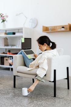 Asian woman working remotely on laptop, sitting in armchair with coffee, in modern home office.