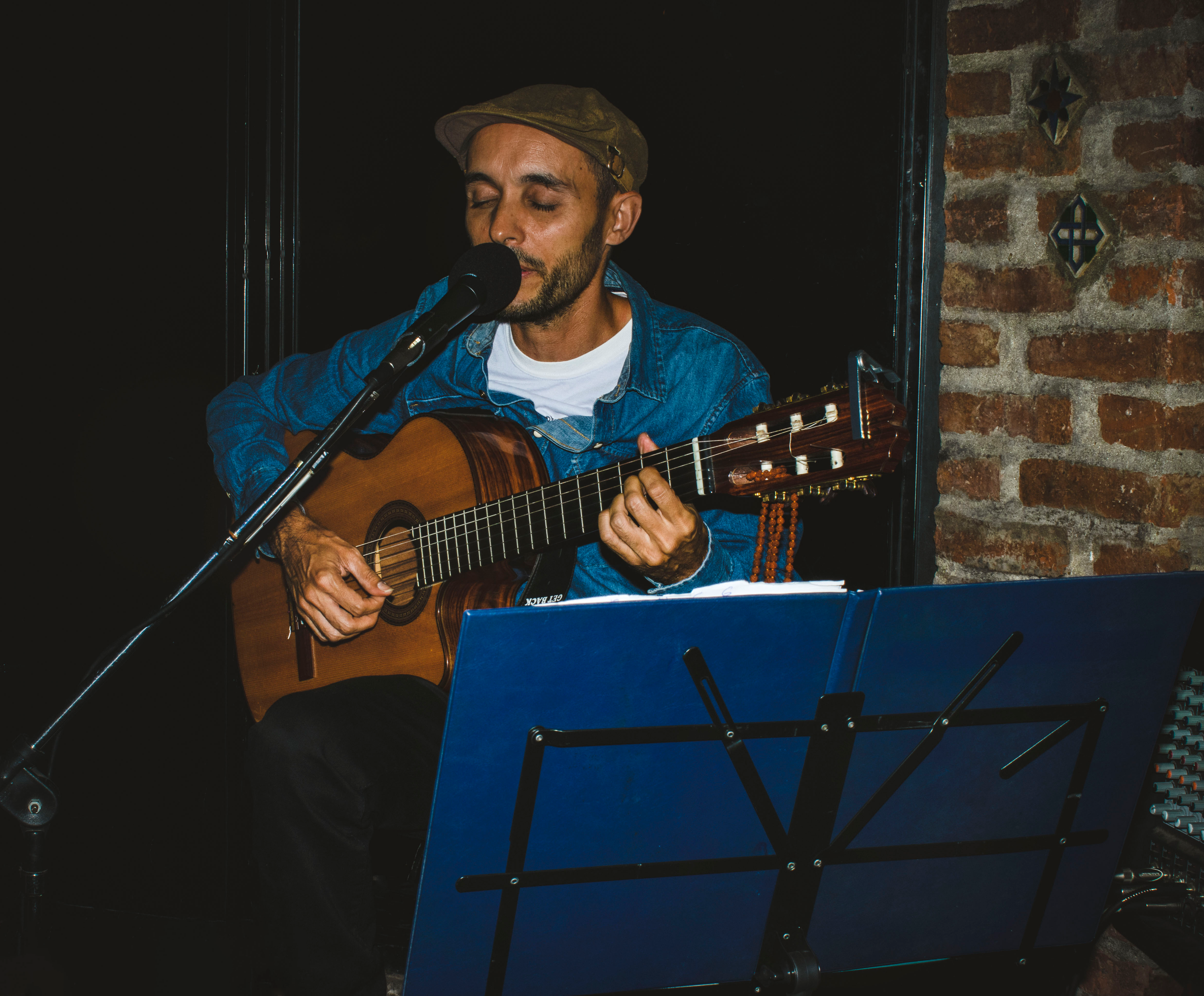 Musician playing guitar and singing into microphone during indoor performance. Rustic background setting.