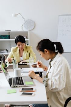 Two Asian women enjoying a lunch break in a contemporary office setting, using gadgets.