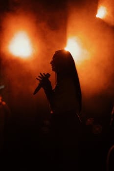 A silhouette of a female performer singing on stage against vibrant orange lights, creating a dramatic atmosphere.