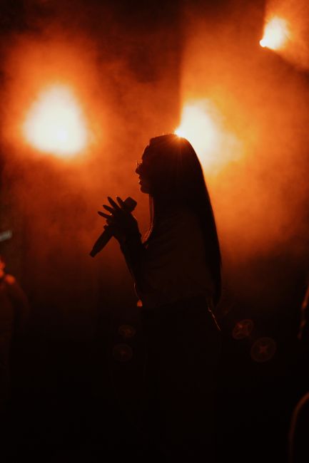A silhouette of a female performer singing on stage against vibrant orange lights, creating a dramatic atmosphere.
