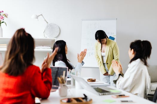 Asian women in an office setting clapping for a colleague after a presentation with a whiteboard in the background.