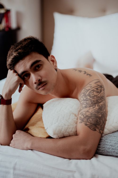 Free Topless man with arm tattoo relaxing on a bed indoors, looking pensively at the camera. Stock Photo
