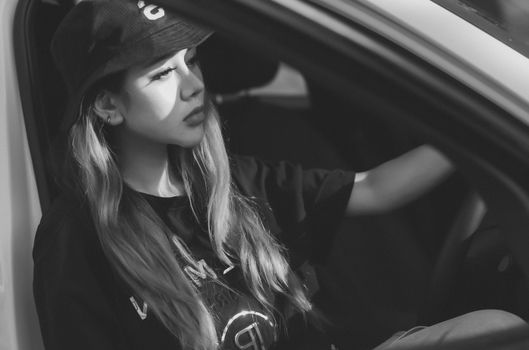 A thoughtful woman in a bucket hat sits inside a white car, captured in black and white.