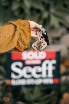 A hand holds keys in front of a blurred sold real estate sign, symbolizing completed sale.