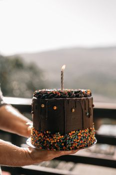 A chocolate cake with sprinkles and candle held outdoors, perfect for celebrations.