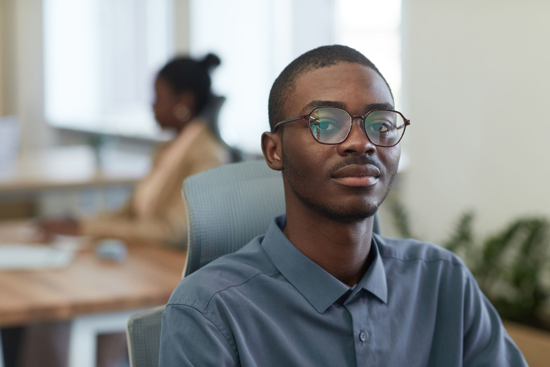 Close-up portrait of a young man wearing glasses, sitting in an office environment.
