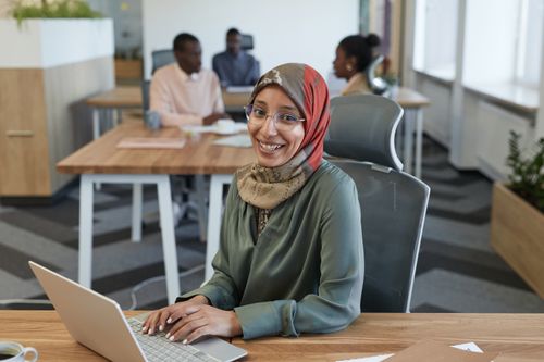Free Smiling woman in hijab working on a laptop in a modern office with peers. Stock Photo