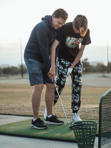 A golf coach crouching beside a junior golfer on a practice green, pointing to a target while the student prepares to chip with a wedge.