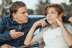 Father expressing frustration while teenage son shows defiance, seated outdoors.