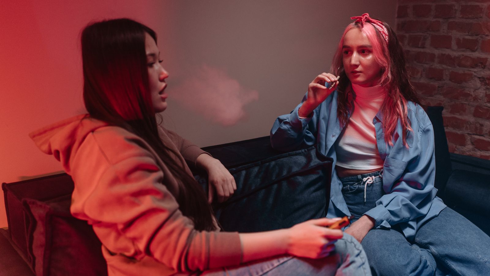 Two women sitting on a sofa indoors, casually smoking and chatting in a relaxed setting.