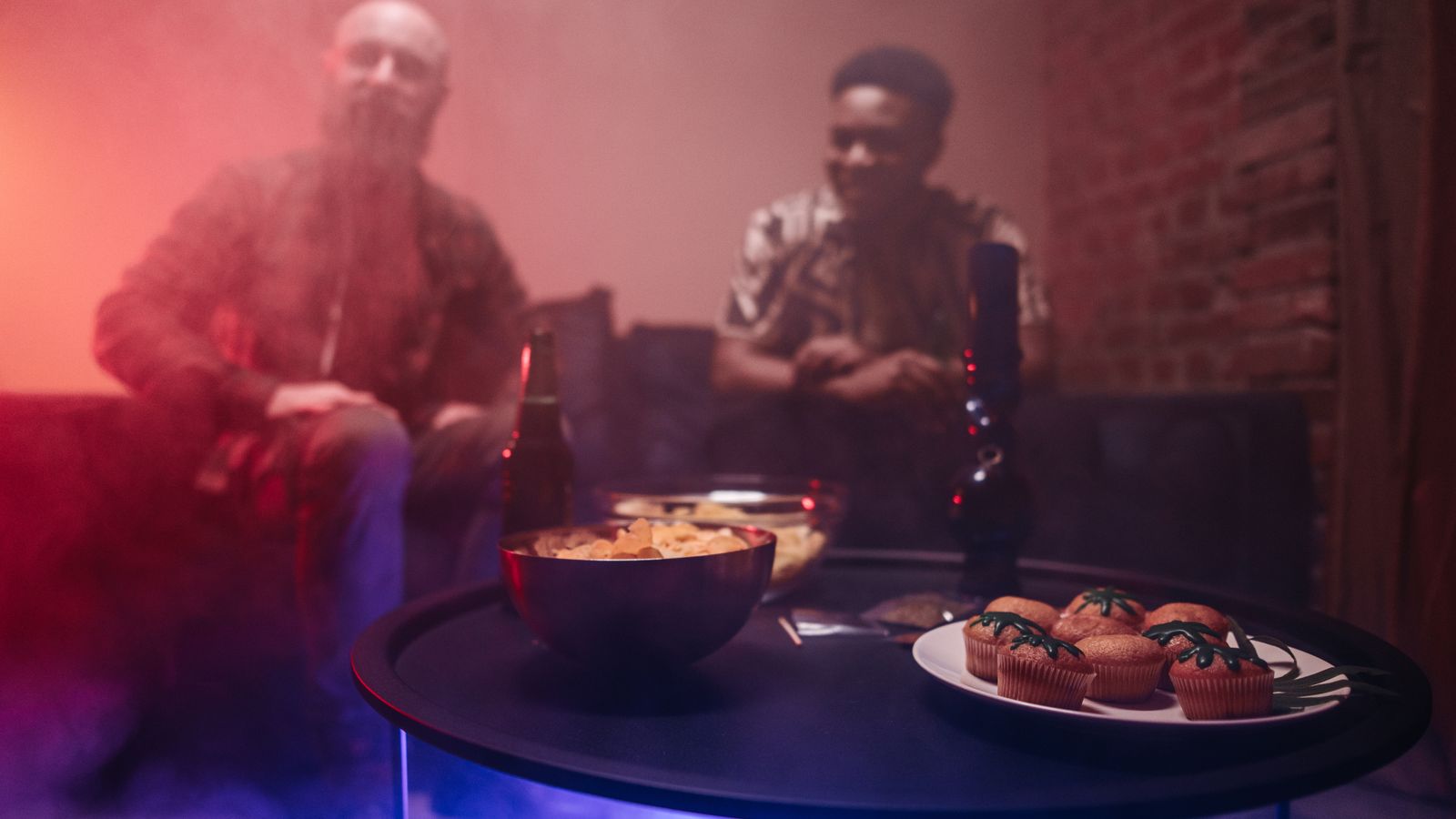 Two people in a dimly lit room with smoke, enjoying drinks and snacks on a table.