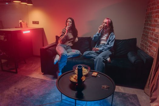 Two women enjoying a relaxed evening with snacks and smoke on a sofa indoors.