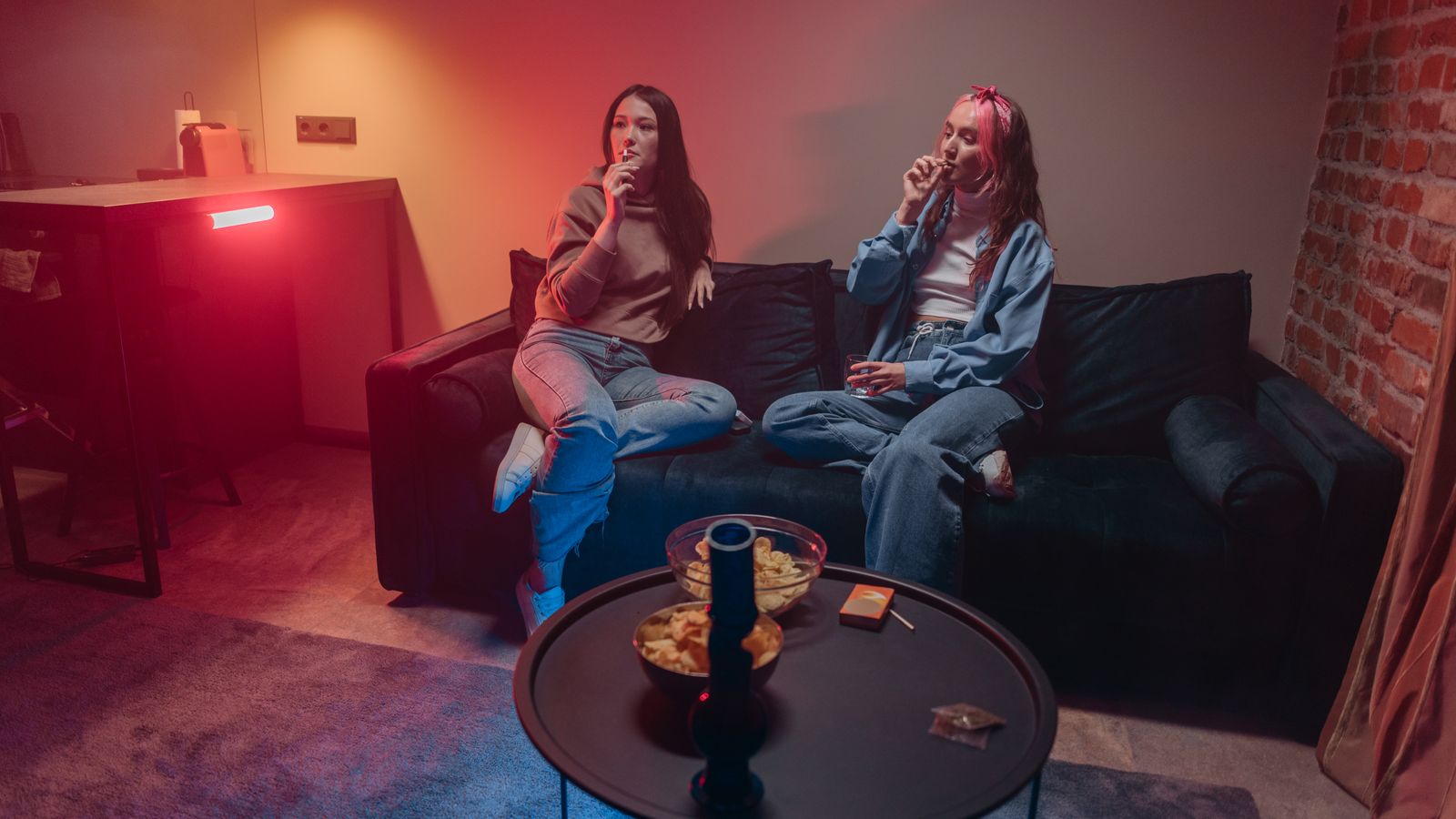 Two women enjoying a relaxed evening with snacks and smoke on a sofa indoors.