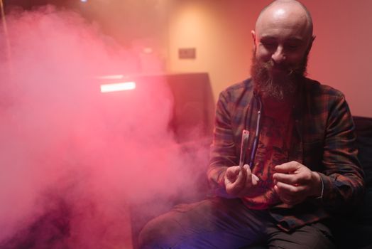Bald bearded man sitting indoors, smiling and enjoying smoke in a relaxed setting.