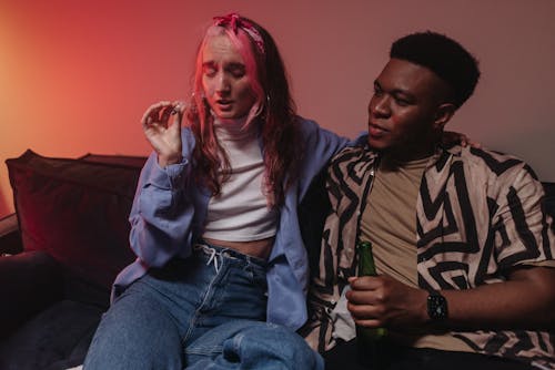 Man in Striped Long Sleeve Shirt Holding Beer Bottle Beside Woman Smoking Marijuana