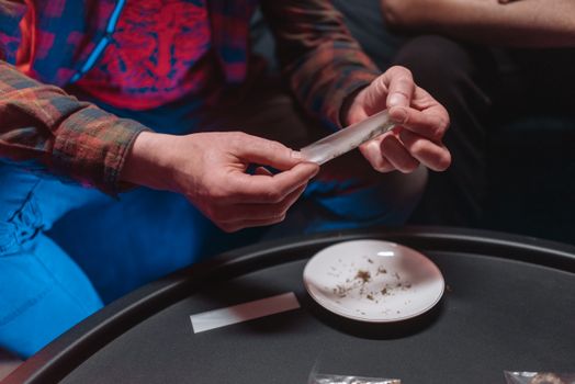 Close-up of hands rolling a joint with cannabis on a plate, capturing a casual scene.