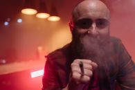 Bald bearded man in checkered shirt smoking indoors under red lighting.