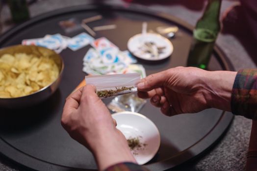 Hands rolling a cannabis joint at a party with snacks and games on a table.