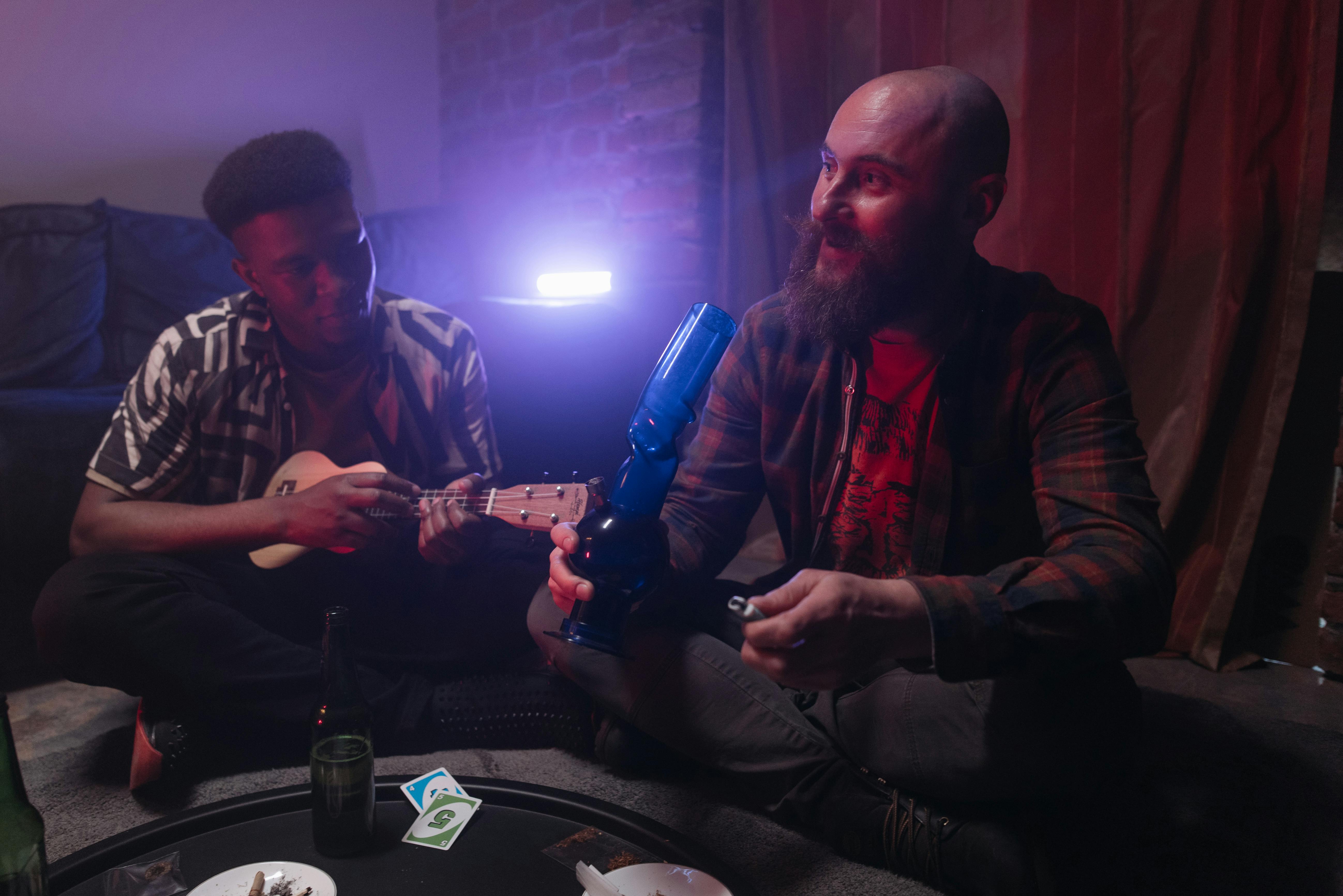 man in plaid dress shirt holding blue glass mini bong beside a man playing ukelele