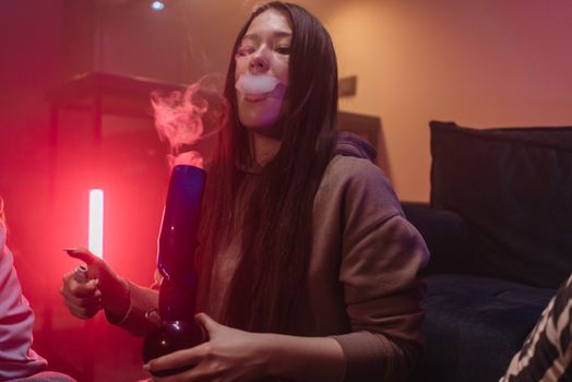A young woman smokes with a bong in a dimly lit room featuring neon lighting.