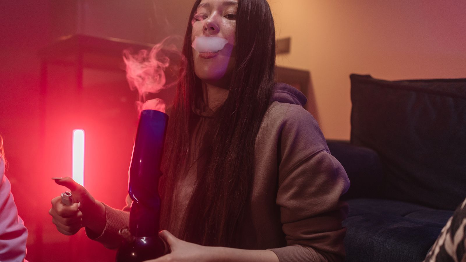 A young woman smokes with a bong in a dimly lit room featuring neon lighting.