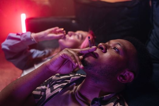 Two friends relaxing on a sofa, smoking indoors with atmospheric lighting.