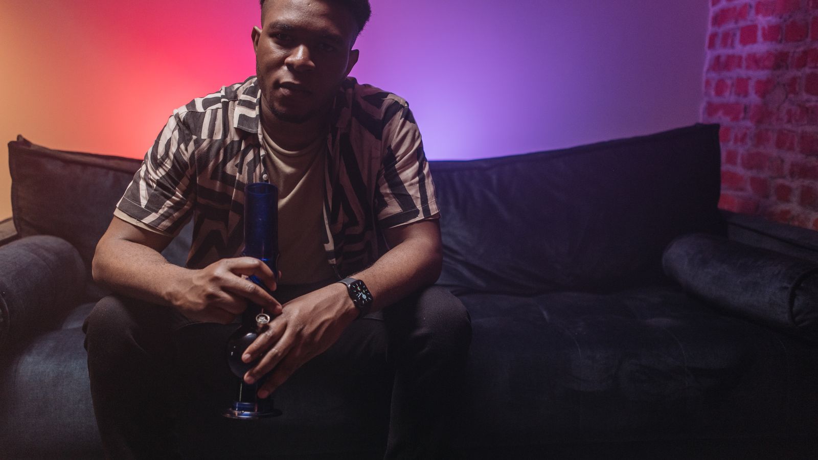 A young man sits on a couch with a bong, ambient colored lights in the background.