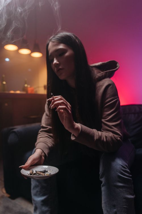 Free A woman in a hoodie sits indoors, smoking, with moody lighting creating a dramatic atmosphere. Stock Photo
