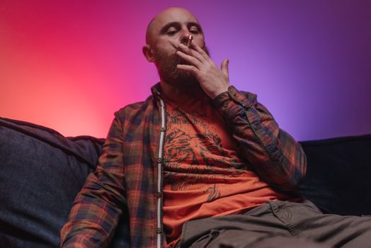 A bearded man smokes indoors against a vibrant, colorful background, creating a relaxing and moody atmosphere.