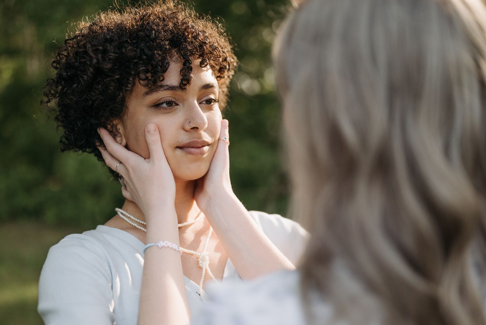 Woman with short curly hair smiling softly, hands gently cupping her jawline outdoors in warm sunlight