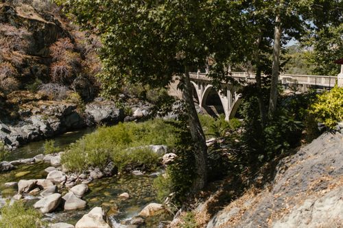 Free Picturesque view of a stone bridge over a flowing river surrounded by lush greenery and rocks. Stock Photo