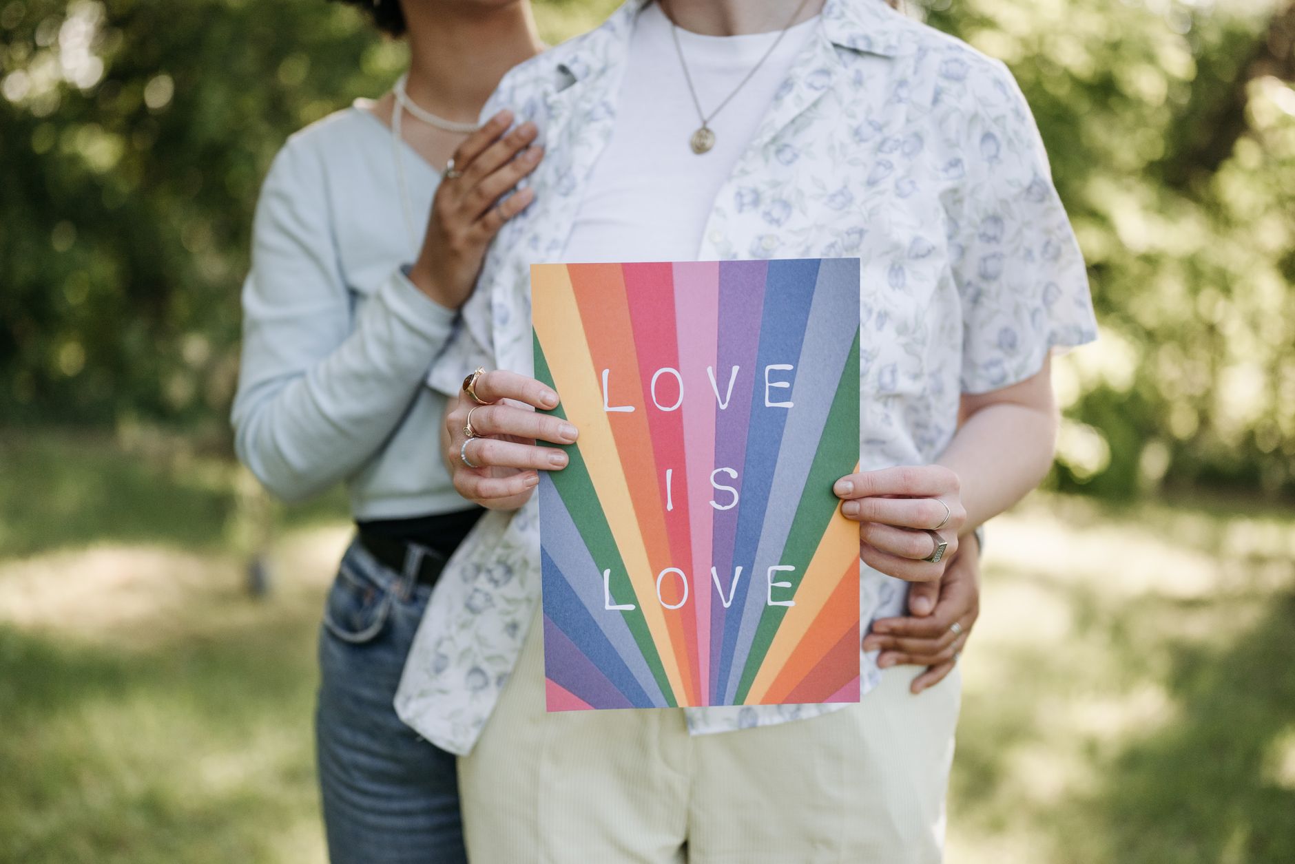 A couple embraces while holding a rainbow poster with 'Love is Love' text in a sunny park.
