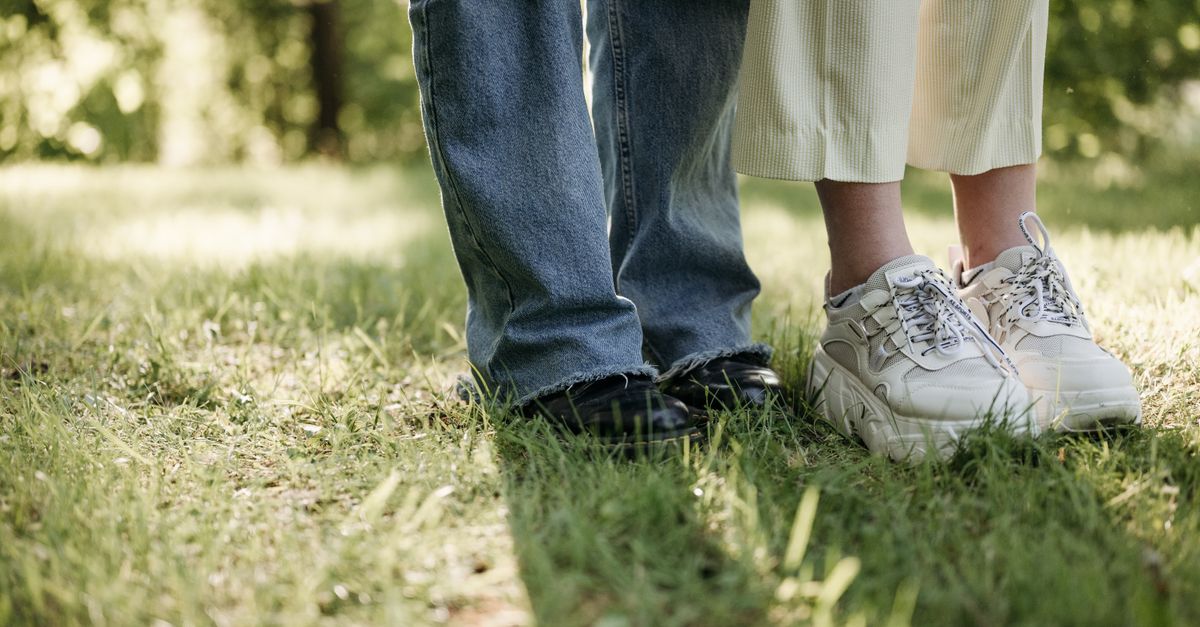 Close-up of two people's feet in jeans and sneakers standing on grass in a sunny park.