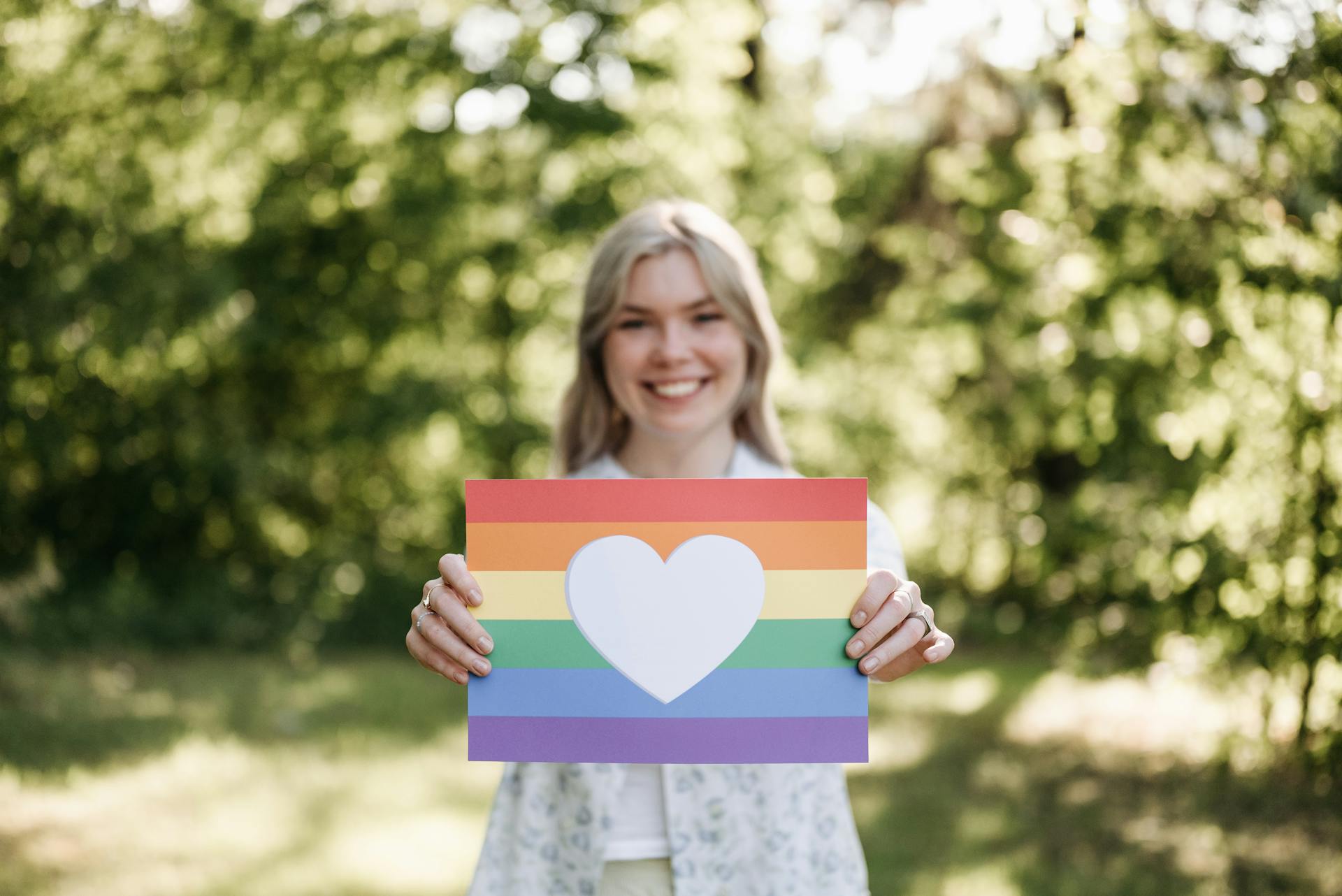 A cheerful woman holds a rainbow pride flag with a heart symbol outdoors, embracing LGBT pride.