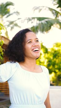 Cheerful woman with curly hair enjoying sunny day outdoors, surrounded by greenery.