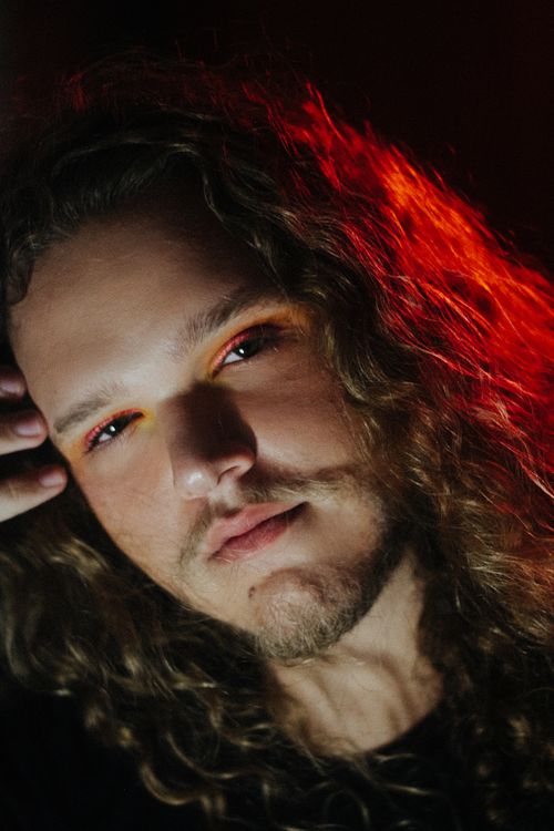 Free Close-up portrait of a man with long curly hair and eye makeup, under warm lighting. Stock Photo
