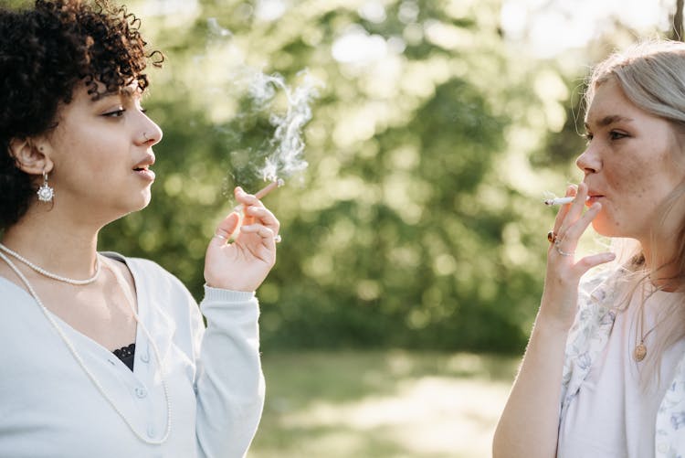 Two Young Women Smoking Cigarettes In A Park 