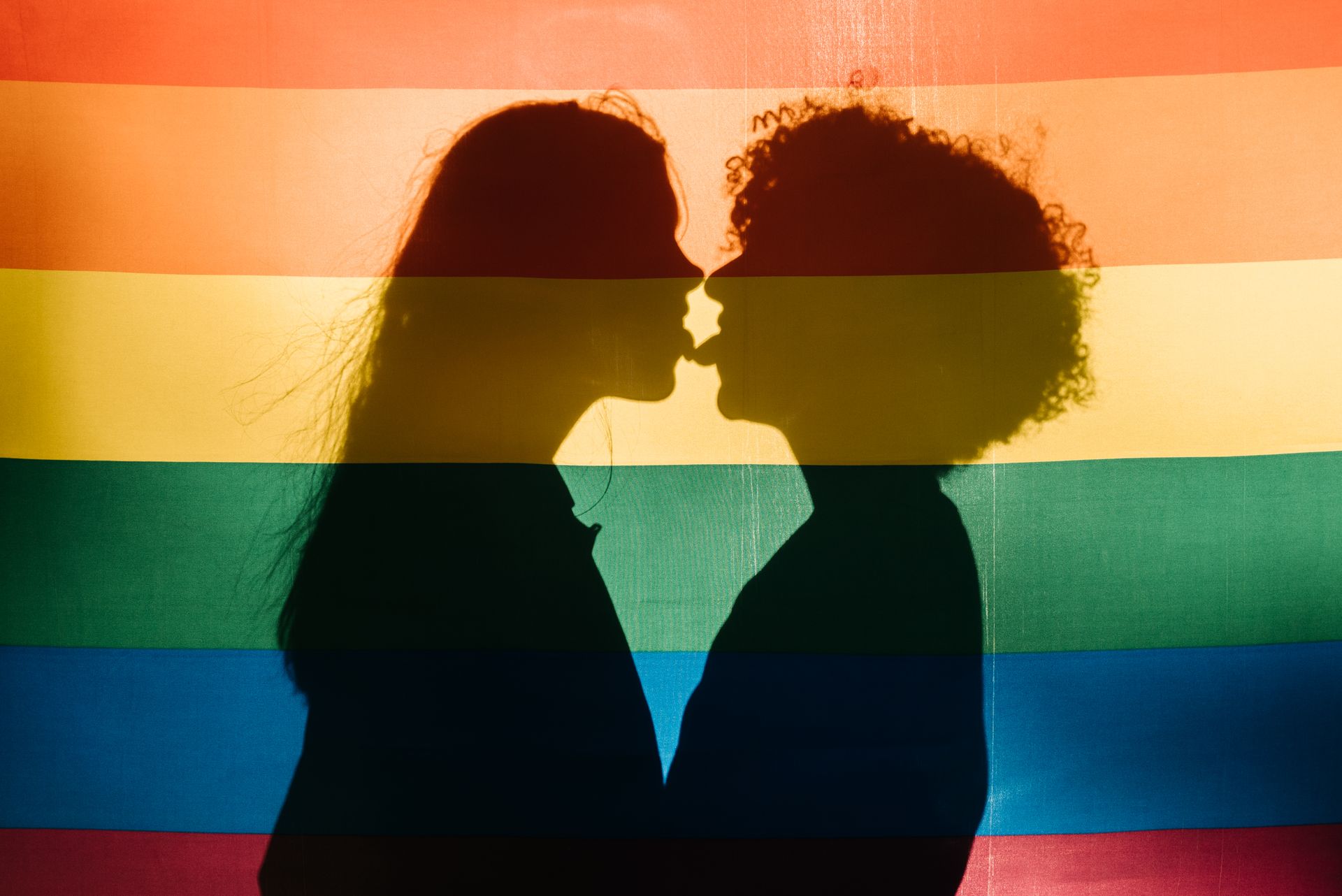 Silhouette of couples showing affection behind a colorful LGBT rainbow flag, capturing love and pride.