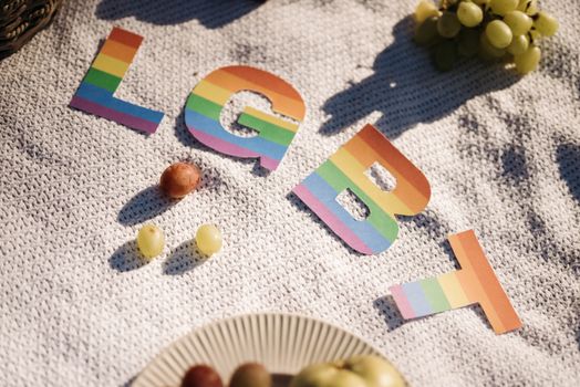 Rainbow-colored LGBT letters on a knit blanket with grapes under sunlight.