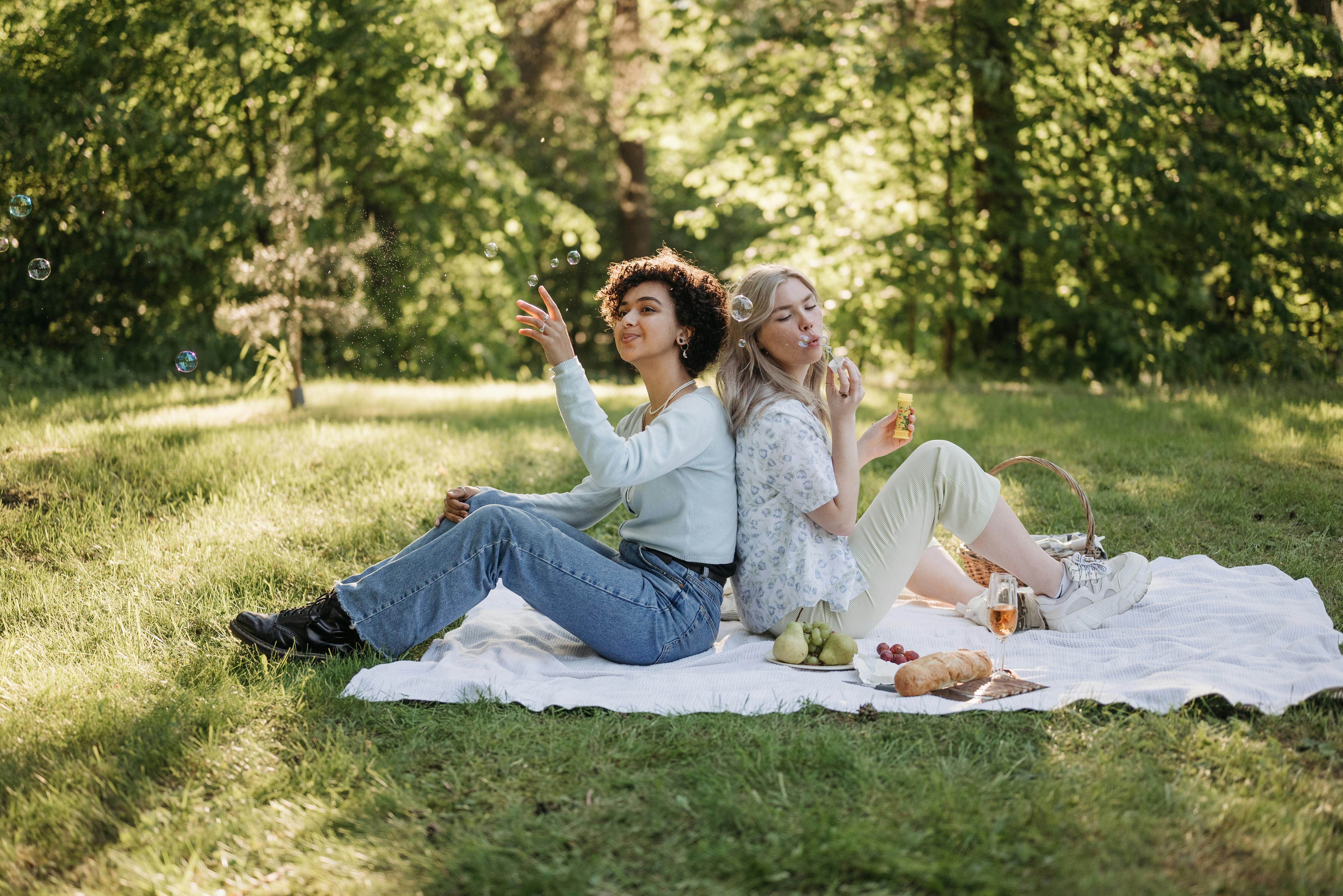 Two women enjoying a sunny picnic in the park, blowing bubbles and relaxing on a blanket.