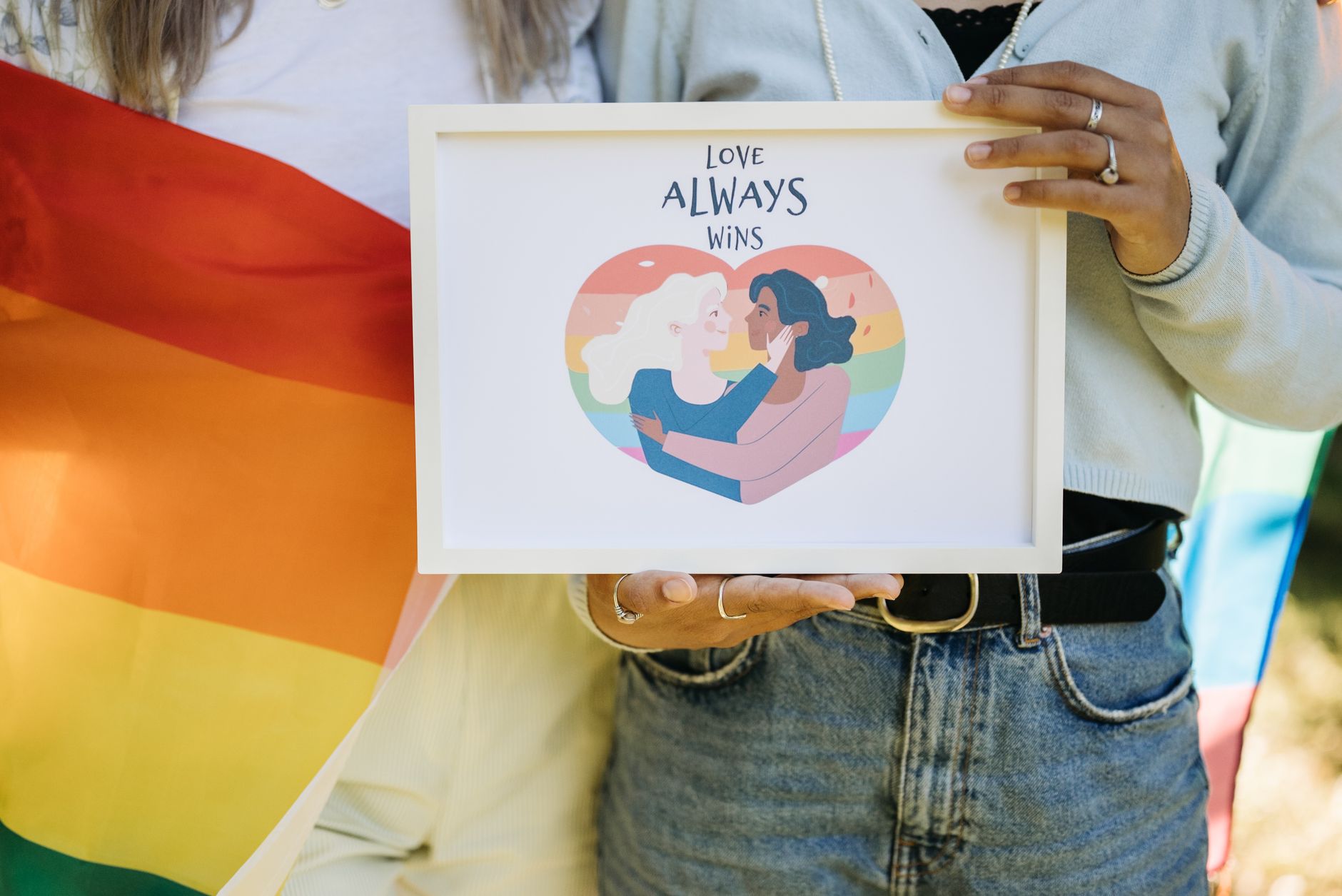Couple holding Love Always Wins sign with rainbow flag, celebrating pride outdoors.