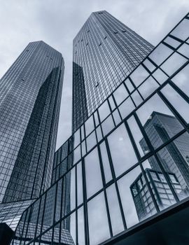 Low-angle view of modern skyscrapers reflecting in a glass facade against a cloudy sky.