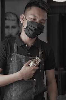 Barber in apron and mask holding hair clipper inside a salon, ready for work.