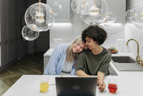 Free Two women sharing a moment of affection in a stylish kitchen with unique lighting design. Stock Photo