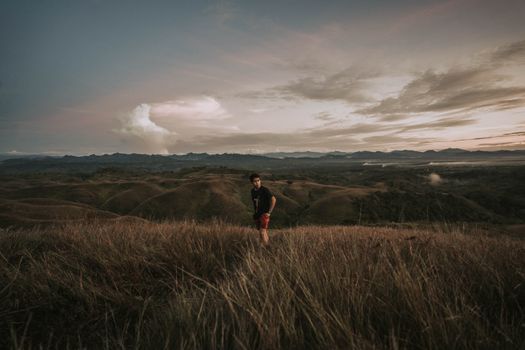 A man in a t-shirt and shorts stands in an expansive grassland during a cloudy sunset.