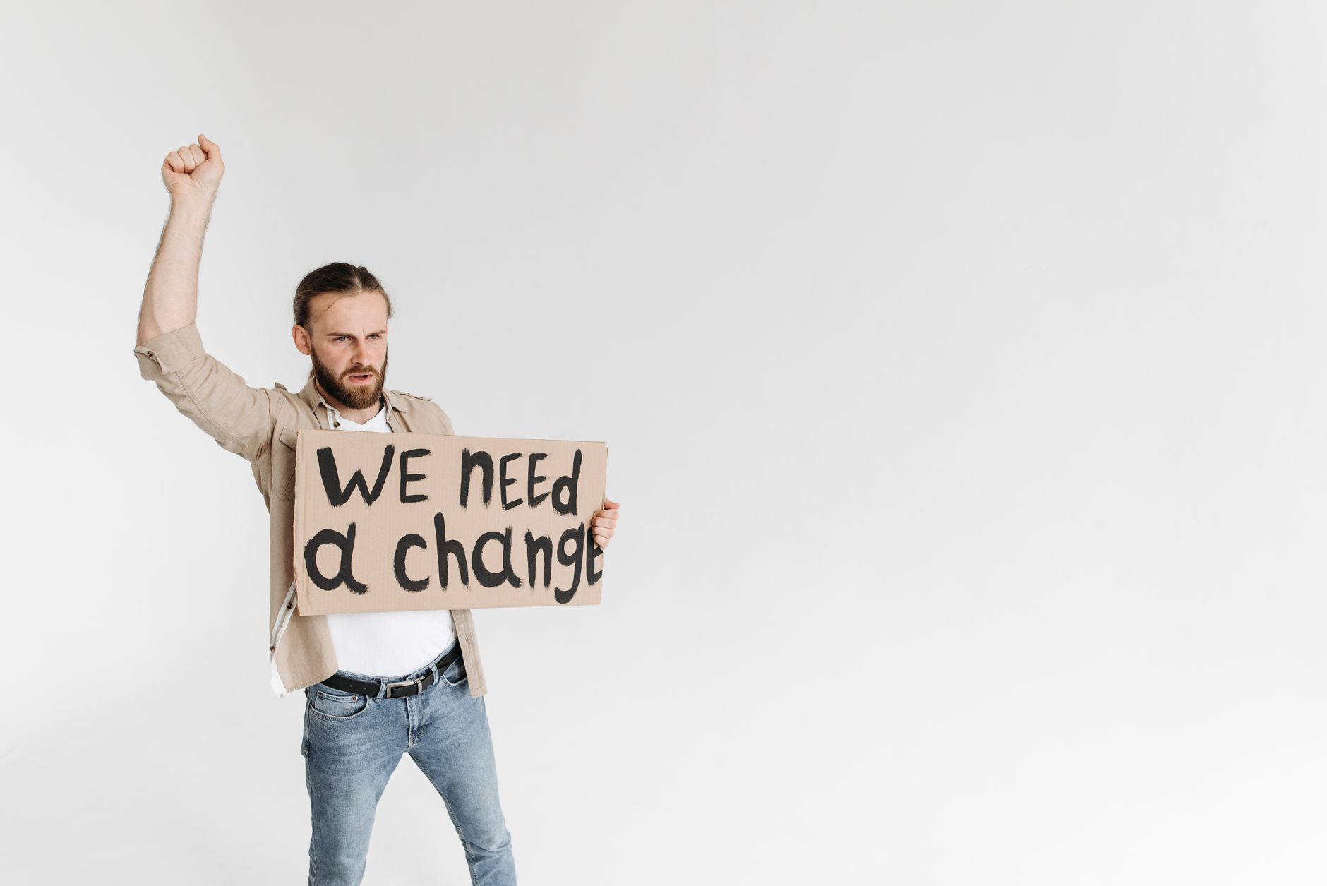 Man holding a 'We Need a Change' sign with raised fist, isolated background. (Photo by Pavel Danilyuk on Pexels)