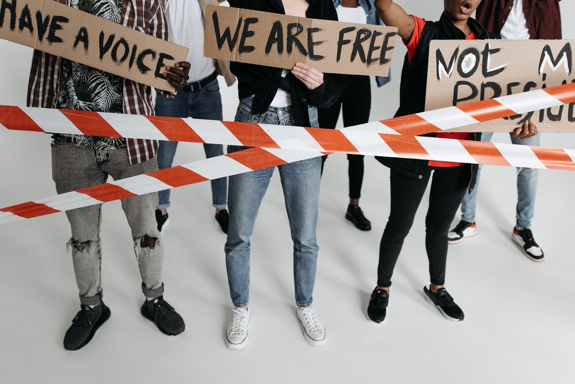 A diverse group of adults holding protest signs behind tape in a studio setting.