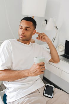 Casual man in white t-shirt holding coffee cup, seated at desk with smartphone.